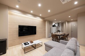 A modern living room with a flat-screen TV, grey sofas, a coffee table, and a dining area with chairs and a table.