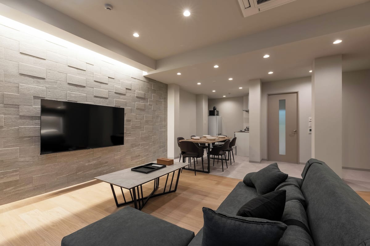 A modern living room with a flat-screen TV mounted on a textured wall, a dining area with a wooden table and chairs, and a gray sofa with cushions.