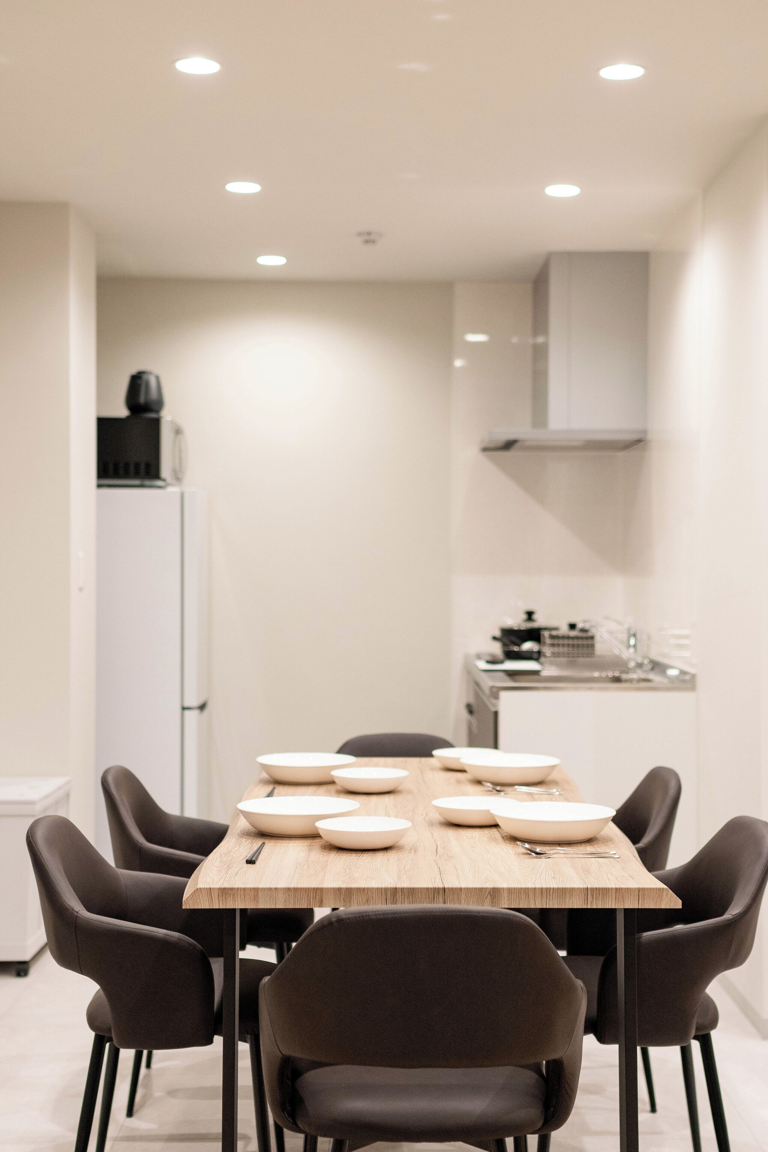 A modern dining area with a wooden table and black chairs, a white refrigerator, and a kitchen area with a stove and sink.