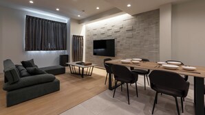 A modern living room with a sectional sofa, a dining table with chairs, and a flat-screen TV mounted on a stone wall. - Crice Hotel Shinsaibashi (Osaka)