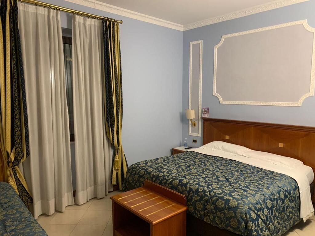 Een hotelkamer met een bed, een nachtkastje, een houten bankje, een raam met gordijnen, en een muur met een spiegel en een fotolijst.