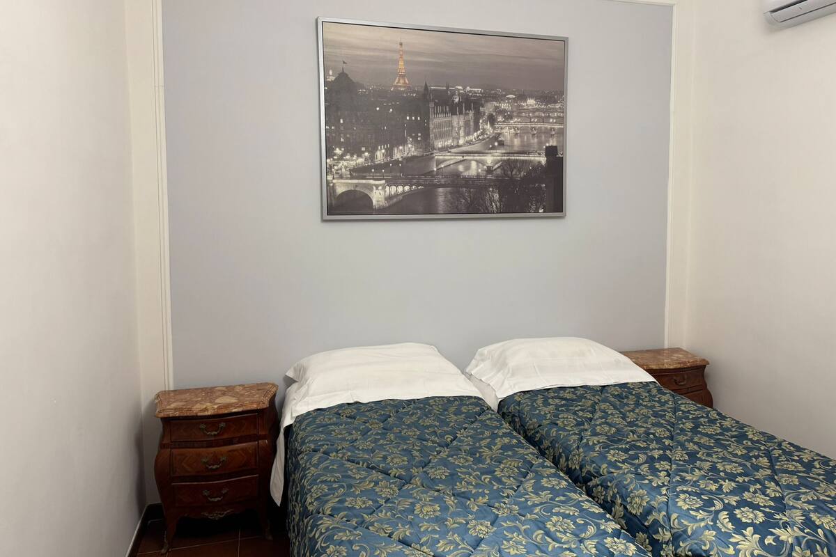 A bedroom with a bed, two nightstands, a wall-mounted air conditioner, and a cityscape picture.