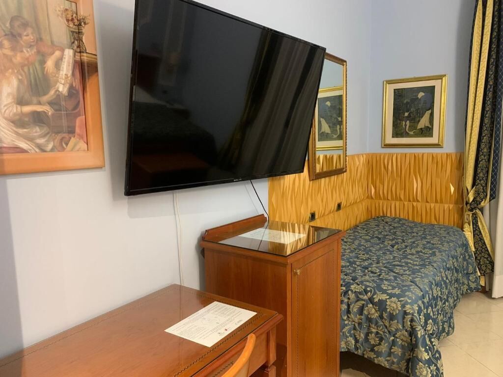 Ein Hotelzimmer mit einem großen Flachbildfernseher an der Wand, einem Holztisch mit Stuhl, einem Bett mit floralem Überwurf und zwei gerahmten Bildern.