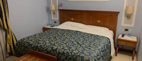Habitación de hotel con una cama, una mesita de noche de madera, una pequeña mesa y una lámpara de pared.
