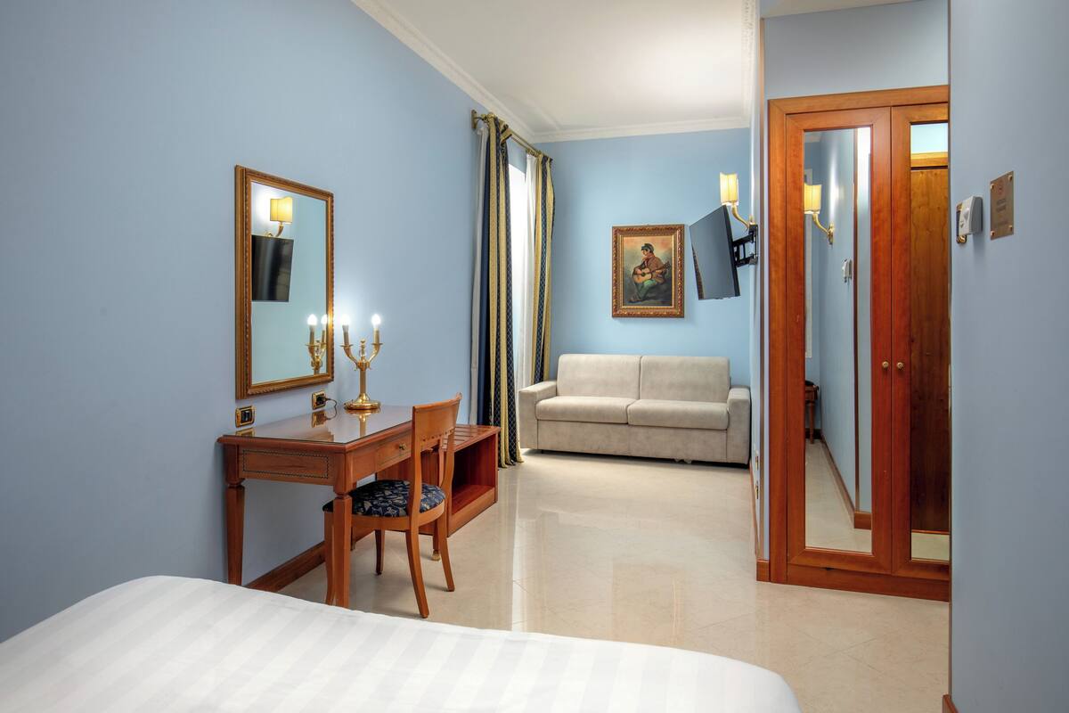 A hotel room with a bed, a desk with a lamp, a chair, a sofa, and a door.
