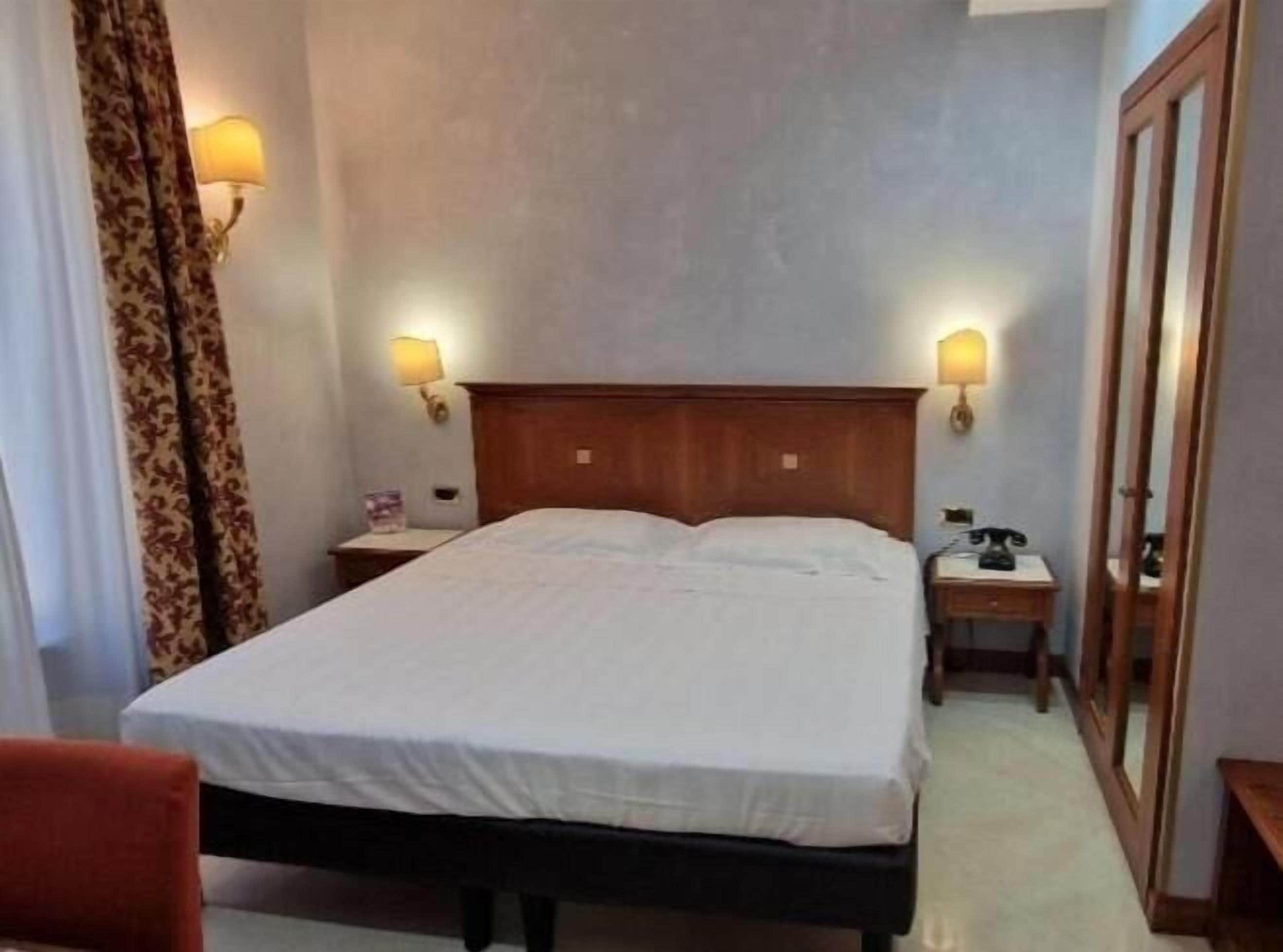 A hotel room with a large bed, two bedside tables, a wooden headboard, and two wall-mounted lamps.