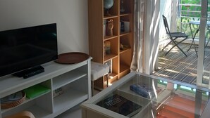 A living room with a TV, bookshelf, and a glass table with books. - Appartement au Calme 10 mn à Pied Centre Ville (La Rochelle)