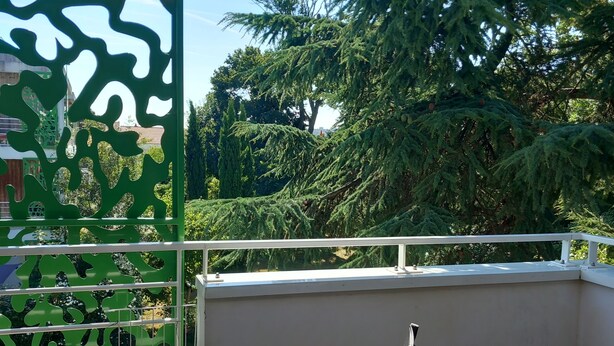A balcony with a table and chairs, a green decorative screen, and a view of trees and buildings. - Appartement au Calme 10 mn à Pied Centre Ville (La Rochelle)