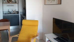 A modern living room with a yellow armchair, a flat-screen TV on a white stand, and a kitchen area in the background. - Appartement au Calme 10 mn à Pied Centre Ville (La Rochelle)