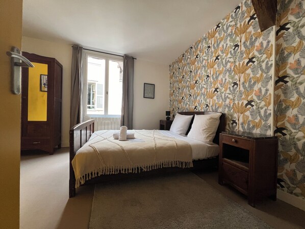 A bedroom with a bed, a nightstand, a wooden wardrobe, a window with curtains, and a wall with a leaf pattern.