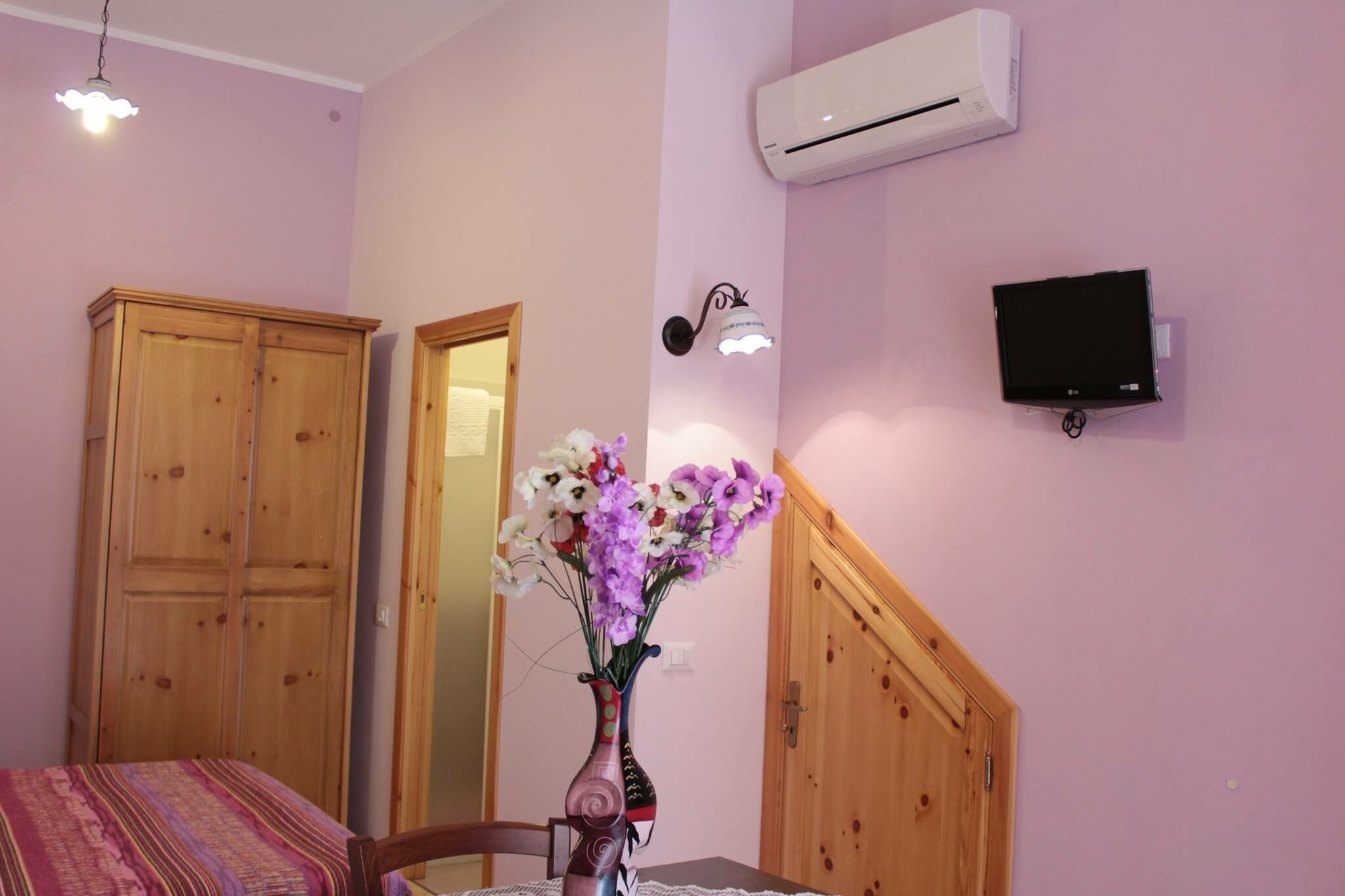 A room with a bed, a wooden wardrobe, a mirror, a vase with flowers, an air conditioner, and a TV mounted on the wall.