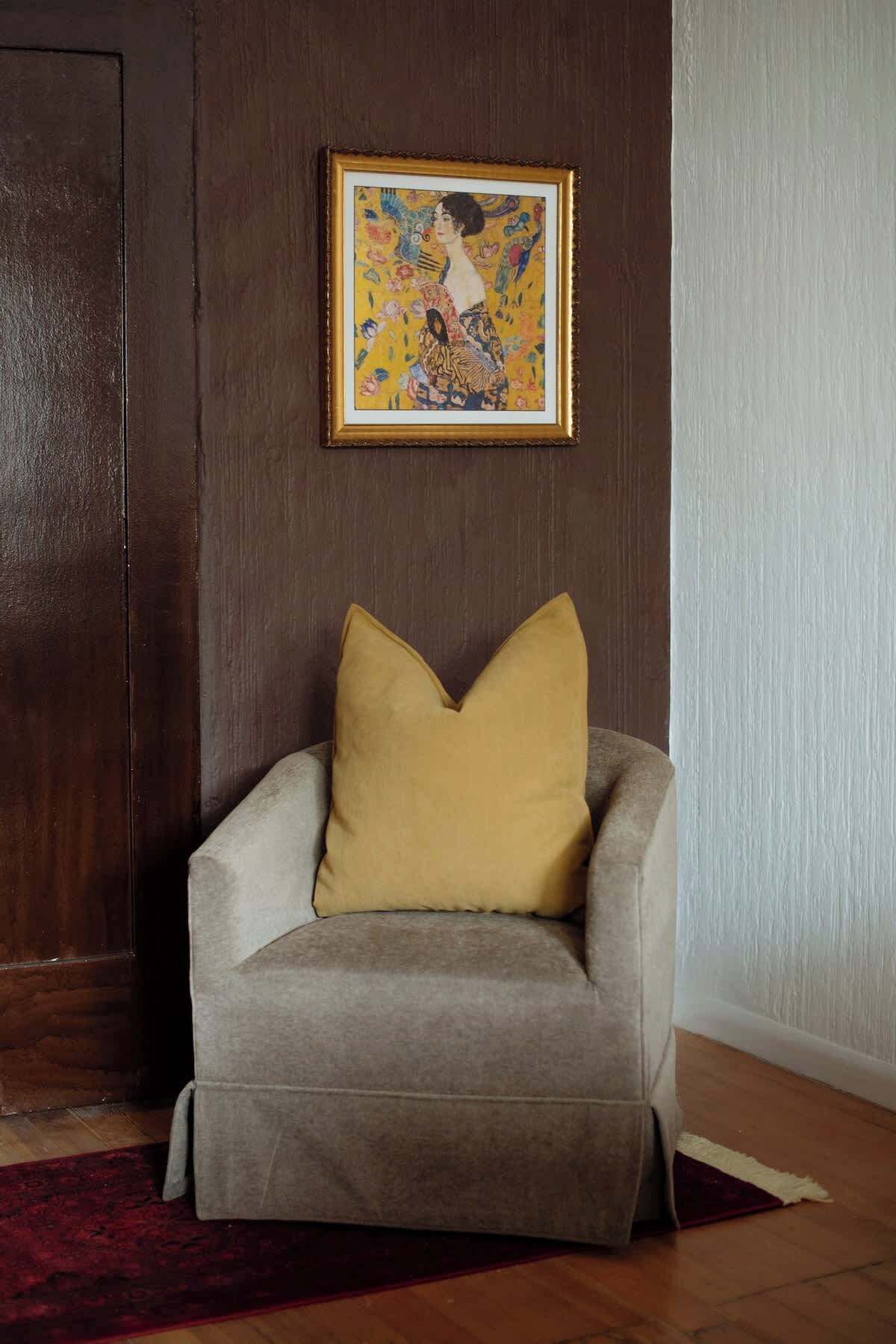 Een grijze fauteuil met een mosterdkleurig kussen tegen een donkere houten wand, met erboven een lijstwerk en een rood tapijt met patroon op de vloer.