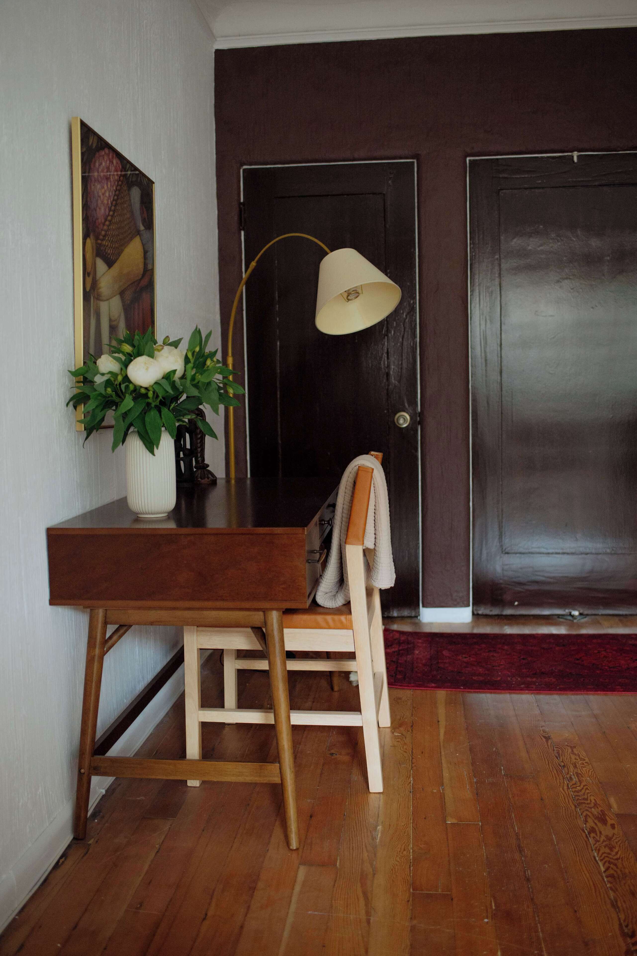A wooden desk with a vase of flowers, a lamp, and a chair in a room with wooden flooring and dark doors.