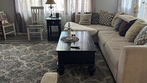 Una sala de estar acogedora con un sofá, una mesa de centro y un sillón. Hay una lámpara sobre una mesita auxiliar y una ventana con cortinas.