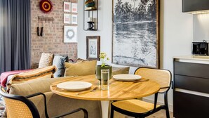 A cozy living area with a round wooden table set for two, a sofa with cushions, a brick wall, and a large framed picture of a beach scene. - Contemporary Charming Apartment The Urban Edge (Cape Town)