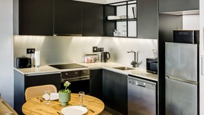 A modern kitchen with dark cabinetry, a stainless steel refrigerator, a microwave, and a dishwasher. There is a wooden dining table with chairs, and a small potted plant on the table. - Contemporary Charming Apartment The Urban Edge (Cape Town)