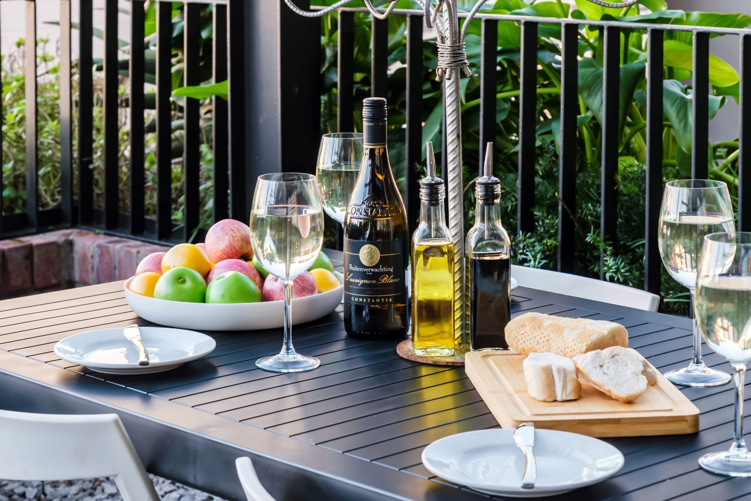 A table set with a bottle of wine, glasses, a bowl of fruit, and a cutting board with bread.