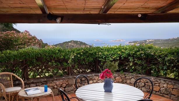 Une terrasse extérieure avec une table et des chaises, un mur en pierre et une vue sur la mer et les collines.