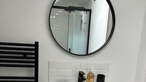 Un baño con lavamanos blanco, espejo redondo, toallero negro y mueble de madera.