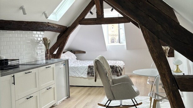 Dormitorio tipo loft con vigas de madera a la vista, pared de ladrillo blanco, cama, mecedora y una pequeña mesa redonda.
