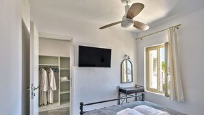 A bedroom with a bed, a ceiling fan, a TV, a mirror, and a window with curtains. - Villa Maria - Naoussa Fishes Blue Grey Suite (Paros)