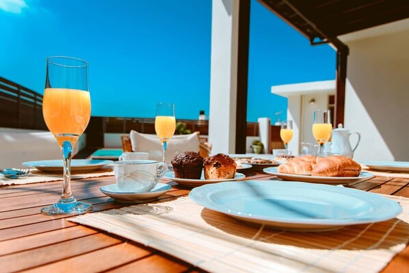 Une table de petit-déjeuner avec des verres de jus d’orange, une tasse, des muffins et des croissants, le tout posé sur une table en bois donnant sur la mer.