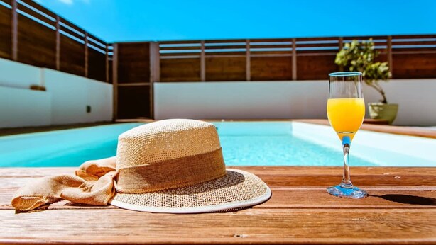 Ein Holztisch mit einem Strohhut und einem Glas Orangensaft am Poolrand.
