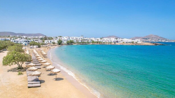 Una playa con aguas azules cristalinas, una hilera de tumbonas y sombrillas, y edificios blancos al fondo.