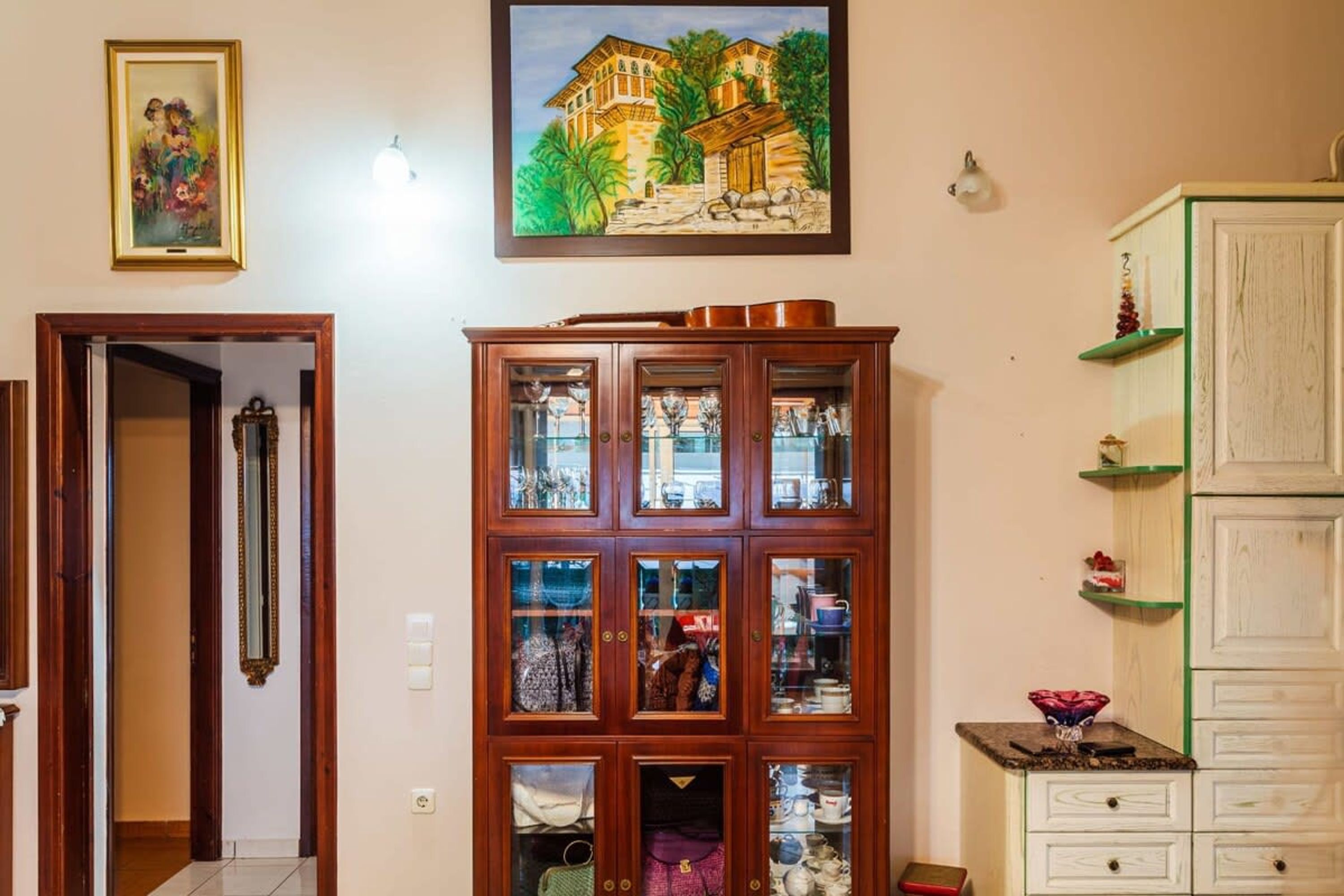 A room with a wooden cabinet with glass doors, a painting on the wall, and a green shelf unit.