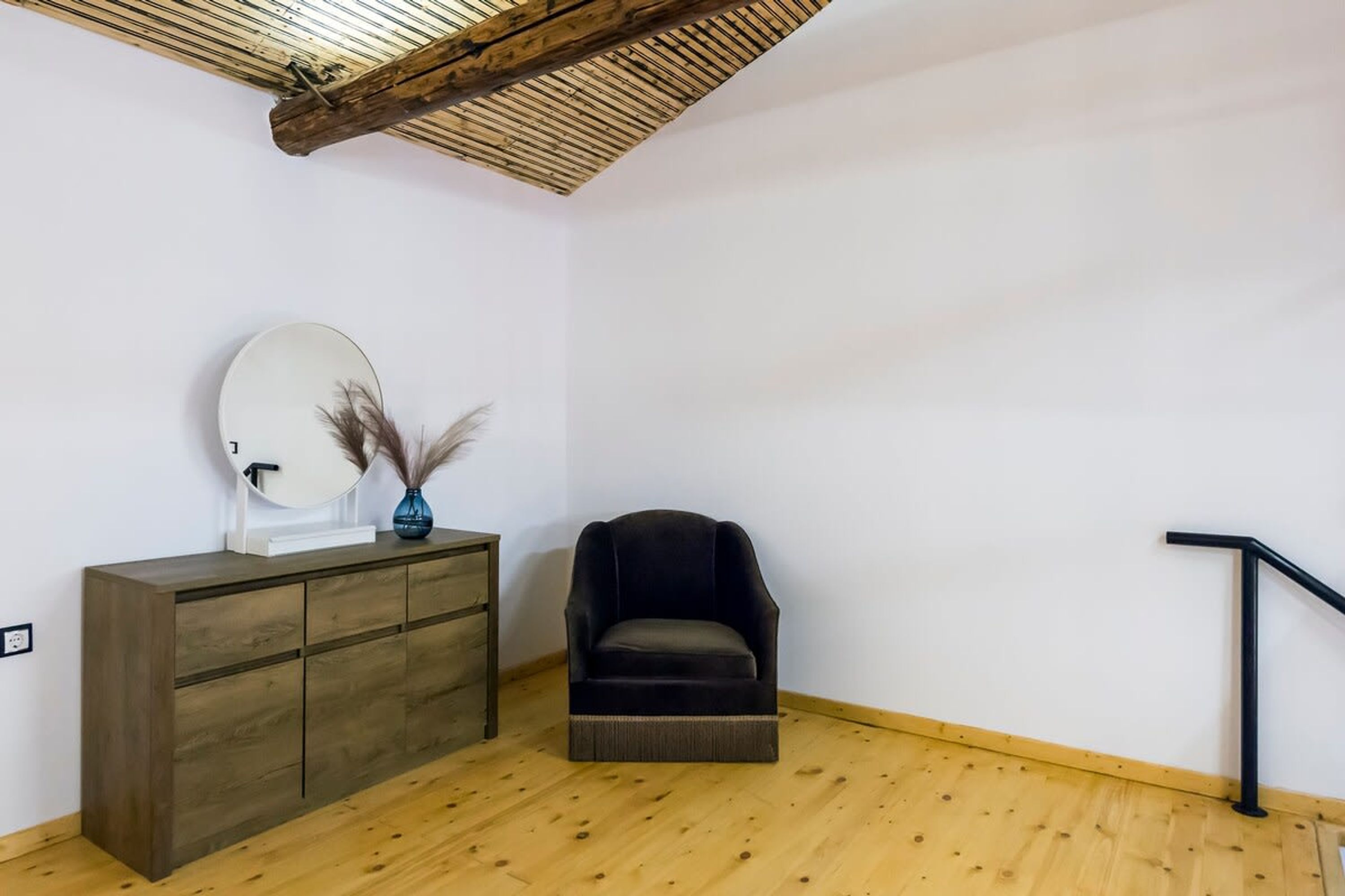 Une pièce avec un plancher en bois, un fauteuil sombre, un meuble en bois doté d’un miroir et une rambarde noire.