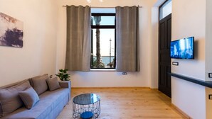 A modern living room with a sofa, a round glass coffee table, a TV on the wall, and a window with curtains. - Aigli Seafront Loft-luxury Design Retreat (Kalamata)