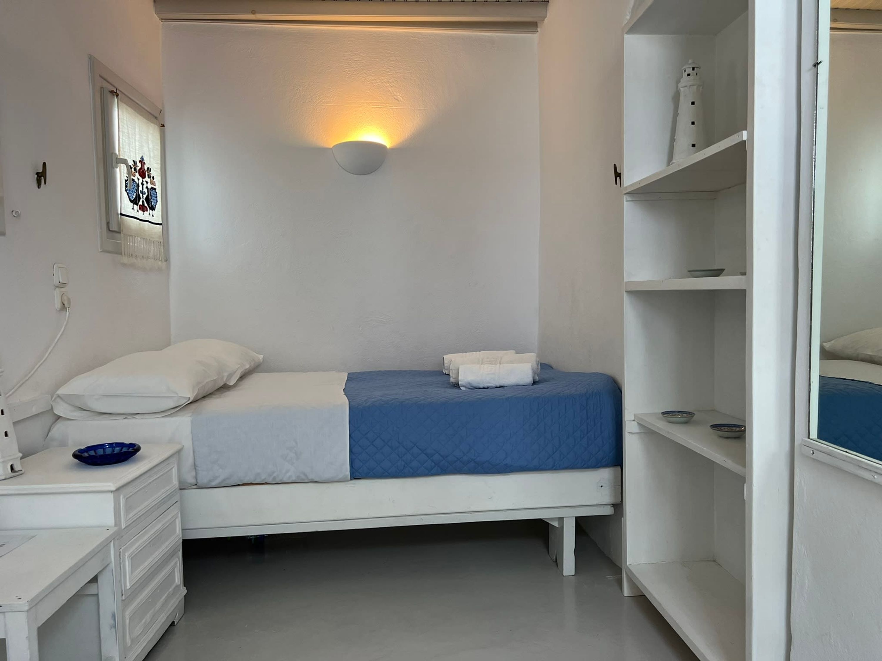 Ein heller Raum mit einem Bett, einem Nachttisch, einer Wandleuchte und einem Regal mit Schalen.