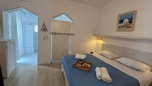 A bedroom with a bed, a bedside table, a nightstand, a window, a door, and a painting on the wall. - Mykonos Violet Nest - Sea View Escape (Mykonos)