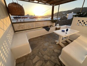 Un espacio de estar al aire libre techado, con suelo de piedra, mobiliario blanco y vistas a barcos y colinas al fondo.