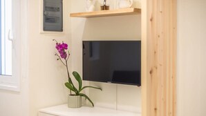 A room with a flat-screen TV mounted on a wall, a small potted plant on a white cabinet, and a wooden shelf above the TV. - The Urban Loft 2 - Athens Unique Oasis (Kaisariani)
