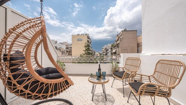 A balcony with wicker furniture, including a hanging chair and two chairs, a small round table with drinks, and a view of buildings and greenery in the background. - The Urban Loft 2 - Athens Unique Oasis (Kaisariani)