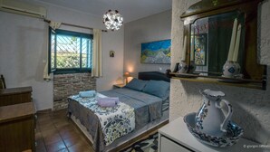 A bedroom with a bed, a nightstand, a dresser, a window with curtains, a hanging light fixture, a painting, and a shelf with decorative items.
