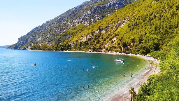 A coastal beach with clear blue waters, a rocky hillside, and a boat in the distance.