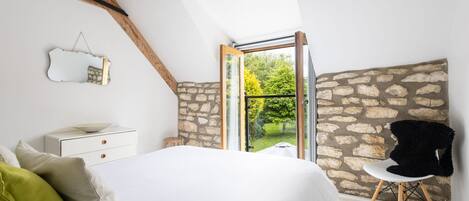 A bedroom with a sloped ceiling, stone wall, a bed with white bedding, a small bedside table, a chair, and a view of greenery through a glass door.