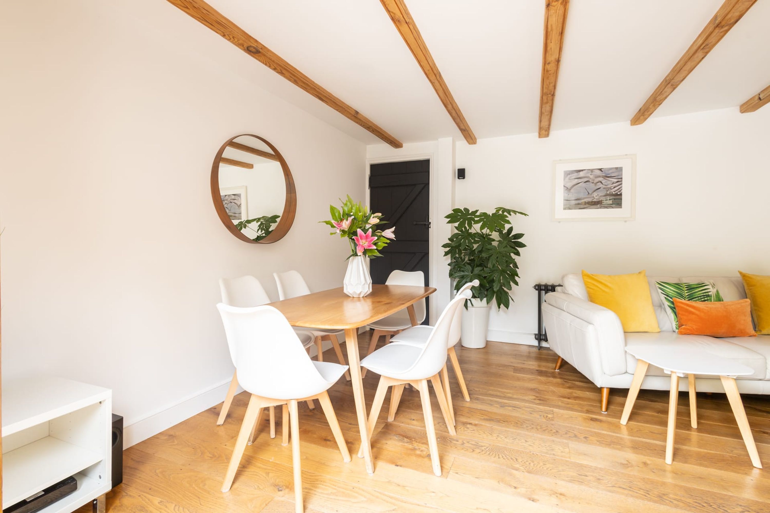 A dining area with a wooden table and white chairs, a potted plant, and a white sofa with colorful cushions.