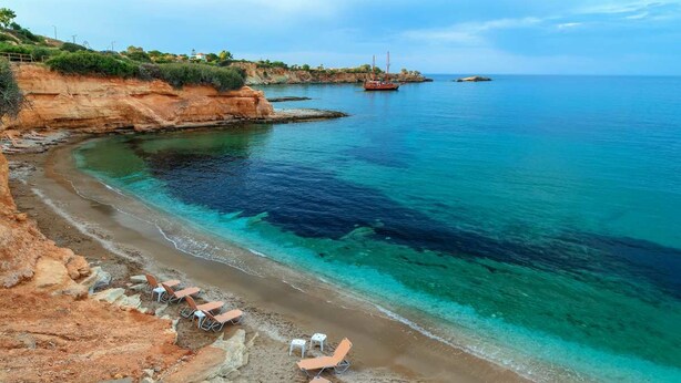 Une plage côtière aux eaux turquoise et limpides, bordée de falaises rocheuses, avec quelques bateaux visibles au loin.