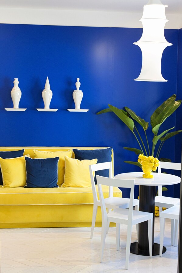 Una habitación con un sofá amarillo, sillas blancas, una mesa redonda y una pared azul adornada con jarrones blancos.