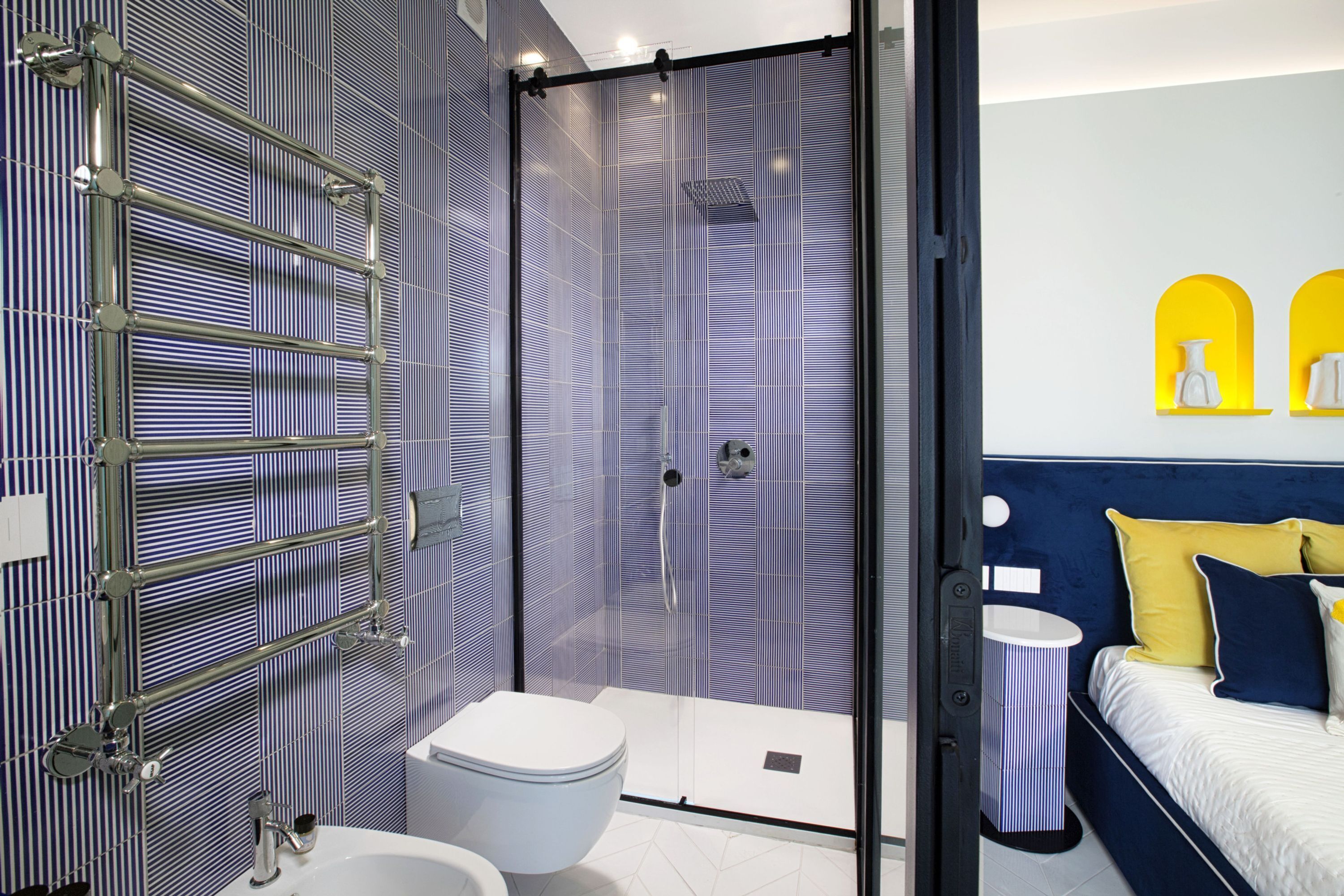 A modern bathroom with a glass shower enclosure, a white toilet, and a bedside table with a lamp.