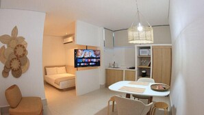 A modern living room with a sofa, a dining table with chairs, a TV mounted on the wall, and a kitchen area with cabinets and a hanging light fixture. - Roka Hotel Boutique (Boa Vista)
