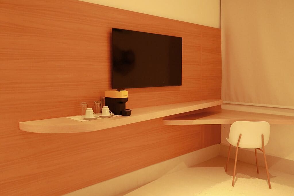 Una habitación moderna con una pared de madera, un televisor montado, una silla blanca y una pequeña mesa con cafetera y tazas.