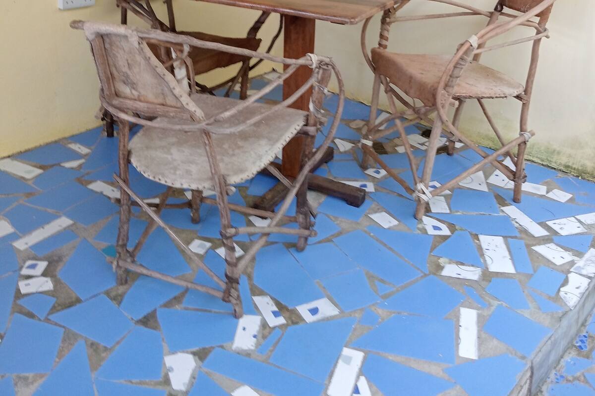 Un pequeño espacio al aire libre con una mesa y sillas de madera, piso de baldosas y una ventana con persianas azules.