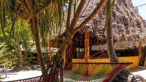 Una hamaca bajo una palmera, una construcción con techo de paja y suelo arenoso.