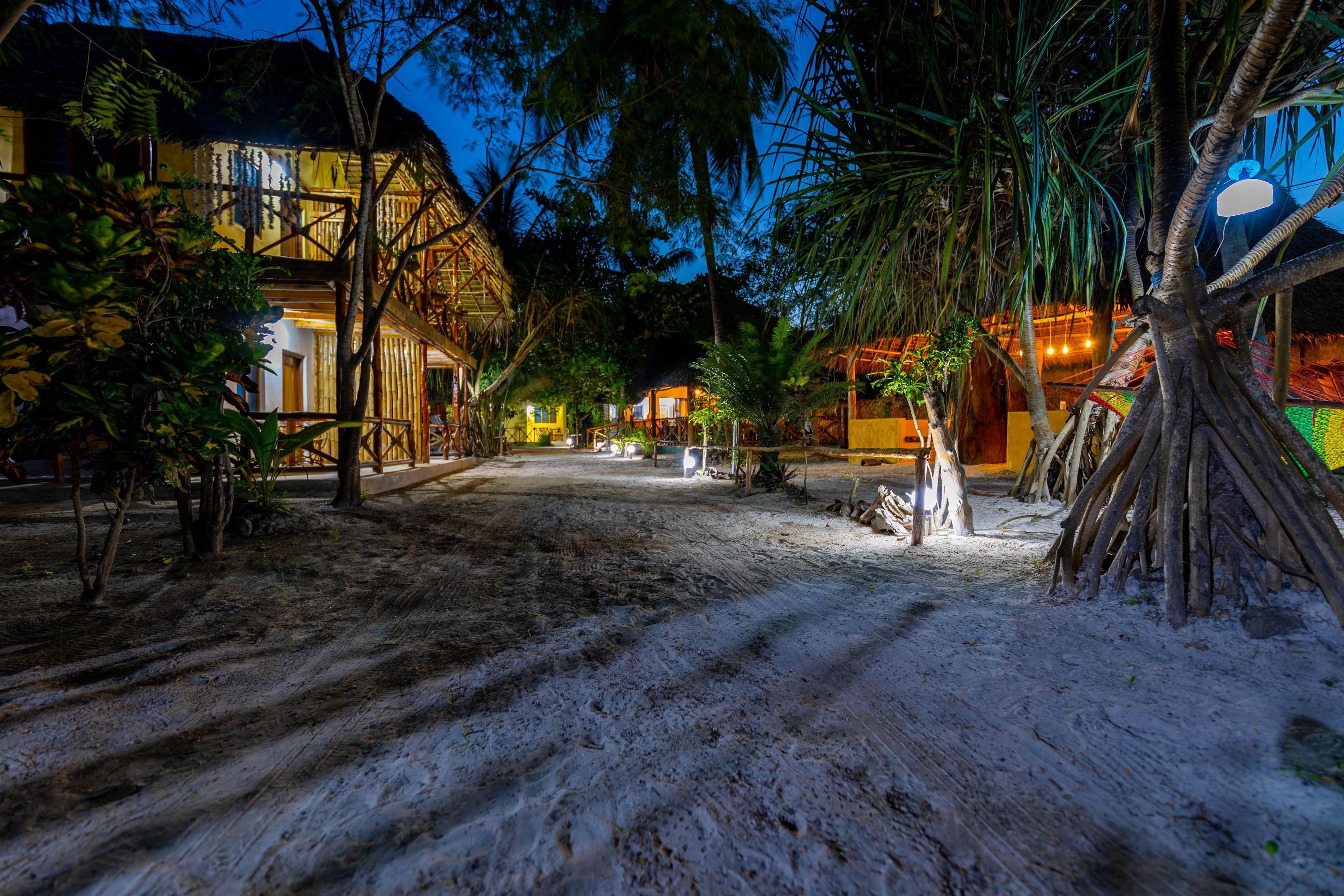 A tropical resort with thatched-roof buildings, a dirt path, and lush vegetation.