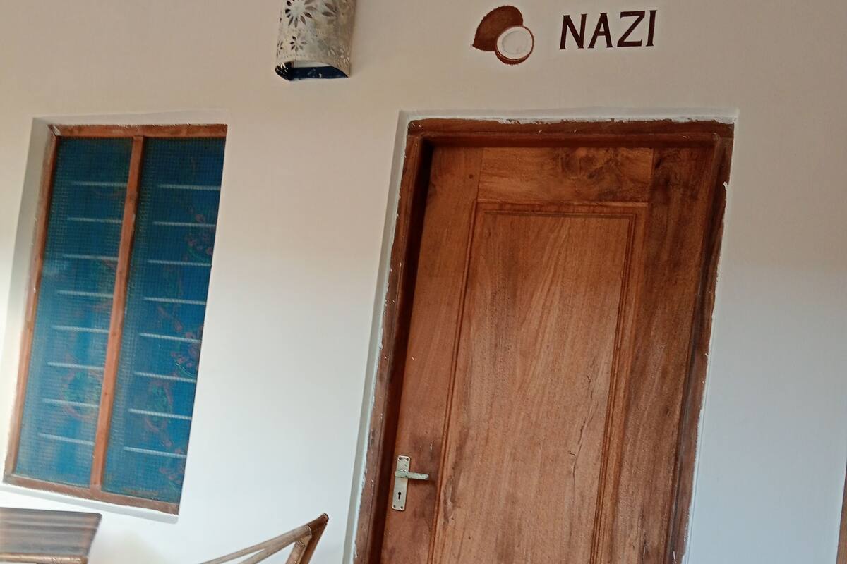 Una puerta de madera con un cerrojo metálico, una silla de mimbre, una ventana con mosquitera y un aplique de pared.
