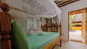 Una cama con sábana verde y cabecera de madera, dosel blanco y vistas a un espacio exterior con estructura de paja.